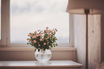 A delicate bouquet of pink roses sits gracefully in a textured vase on a white table. Soft daylight filters through the window, creating a tranquil atmosphere filled with warmth
