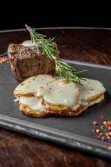 A beautifully plated steak topped with a creamy sauce and garnished with rosemary, accompanied by cheesy potato slices, all set on a dark, elegant serving board.