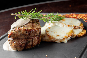 A beautifully plated steak topped with a creamy sauce and garnished with rosemary, accompanied by cheesy potato slices, all set on a dark, elegant serving board.