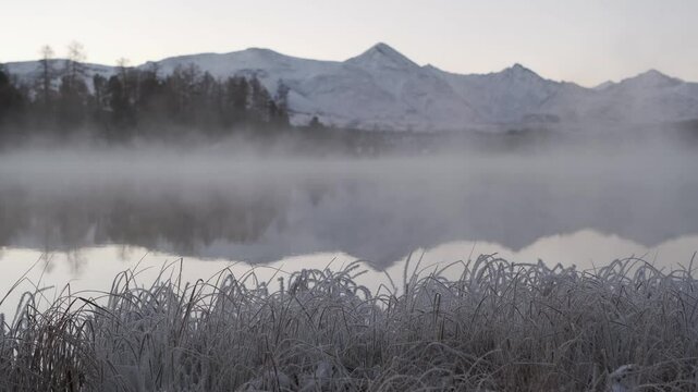 Fog creeps over the unfrozen Kidelu Lake against the background of forest and high snowy mountains at dawn in winter. Amazing Altai landscape