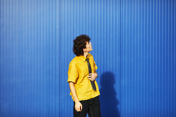 Young student in yellow shirt and tie stands against vibrant blue wall looking up in an urban...
