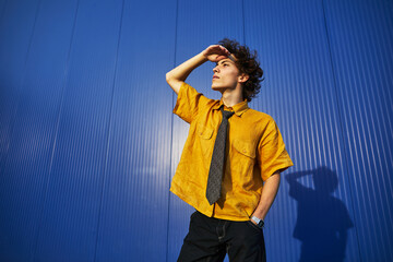 Student in bright yellow shirt and tie posing outdoors against a blue wall in sunlight, showcasing...