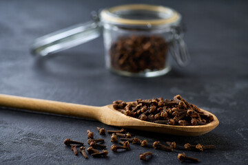 cloves spice in a wooden scoop and in glass storage jar on a black stone table, selective focus.