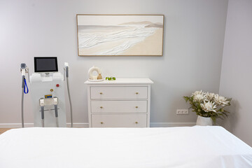 A minimalist room features a white dresser with decor, a medical device with handles, abstract artwork, and a vase of white flowers on the side.
