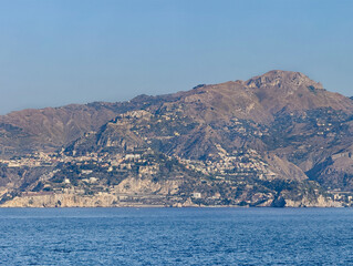 Fototapeta premium Taormina, Sicily, Italy - July 5, 2024: Taormina coastline from Isola Bella with Mendolia beach in center to the rocks of Mazzaro. Cityscape on the flank of the mountain range under blue sky