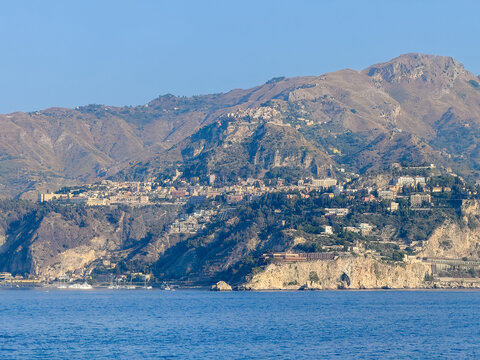 Taormina, Sicily, Italy - July 5, 2024: Taormina cityscape above sea level. Yacht harbor protected by Isola Balle cliffs with the large UNAHOTELS Capotaormina on top. Mountain range and blue sky and s