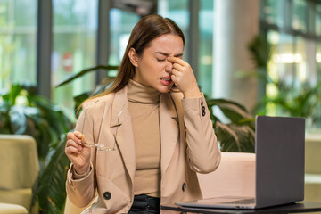 Overworked young Caucasian businesswoman suffering from eye strain and headache working on laptop...