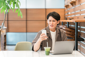 カフェでスマホとパソコンを使う若いアジア人男性（ニュース・驚く・悲しい）
