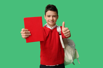 Happy schoolboy with backpack and book showing thumb-up gesture on green background