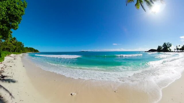 Gentle turquoise waves wash onto the pristine white sand beach with a clear blue sky and distant islands on the horizon. Seychelles, Praslin. Constance Lemuria Beach.