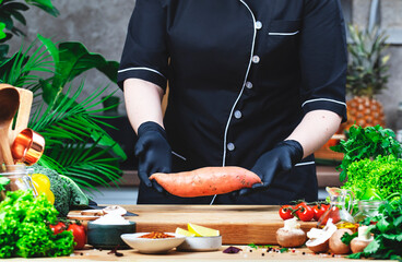 Cook in uniform holds sweet potato.. Cozy kitchen with wooden table, kitchenware, vegetables, herbs and ingredients for cooking. Healthy vegan eating, culinary, recipes, food blogging