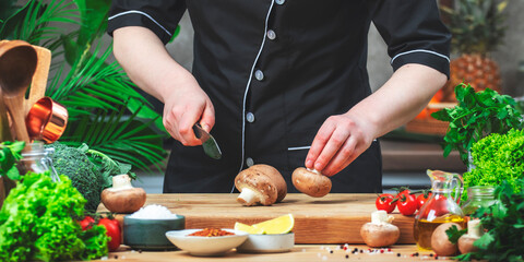 Chef holds fresh brown mushrooms and knife in hands . Cozy kitchen with wooden table, kitchenware, vegetables, herbs and ingredients for cooking. Healthy vegan food, culinary, recipes, food blogging