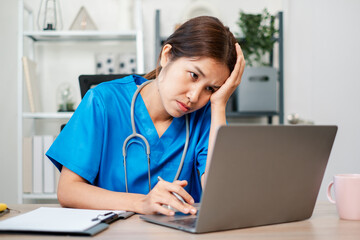 Exhausted nurse in blue scrubs using a laptop, showing stress and fatigue in a healthcare setting.