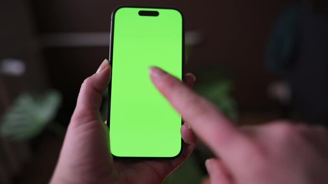 Person Using Smartphone with Green Screen for Mockup Close-up of hands holding tapping smartphone green screen for app design, mockups digital content creation. Blurred indoor background adds depth.