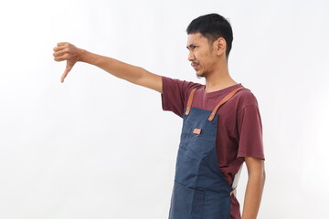 Side view of Discontent Asian man wearing blue apron shows disapproval sign, keeps thumb down,...