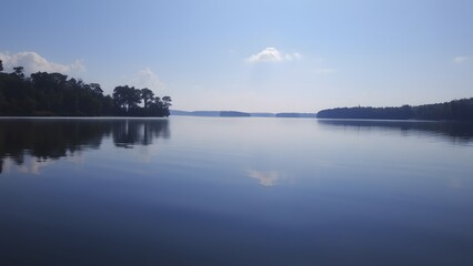 Tranquil lake landscape with crystal-clear water mirroring the vivid blue sky and lush tree-lined shoreline on a sunny day, creating a serene natural atmosphere.