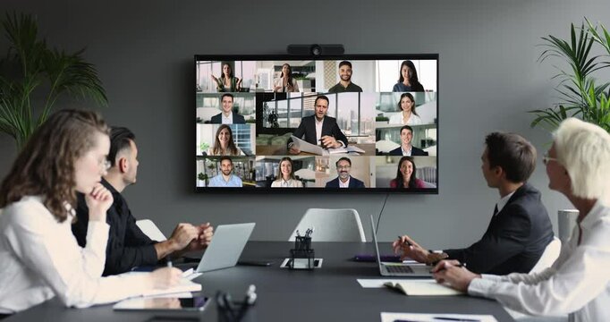 Group of diverse businesspeople gathered in boardroom engaging in video conference with multiple participants displayed on large screen. Global communication and collaboration in interconnected world