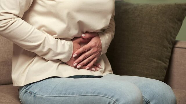 Woman experiencing stomach or abdominal pain, possibly from menstruation, indigestion, or other medical condition, sitting on a sofa and holding her belly
