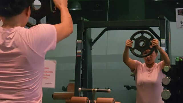 Back view of 40 year old woman doing weightlifting in gym using dumbbells with blurry reflection in mirror.