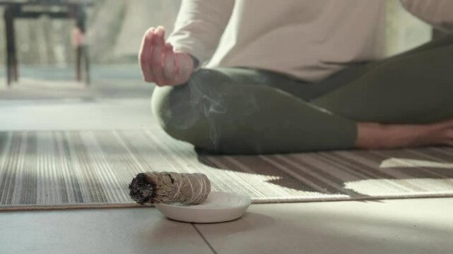 Sunlight illuminates a woman practicing yoga and meditation with her hands in a mudra position, while a smoldering white sage bundle rests on a small plate in front of her