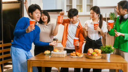 Five young Asian men and women gather around a kitchen desk, enjoying a party with drinks, fried chicken, and bread. They share laughter, good food, and happiness in a lively atmosphere.