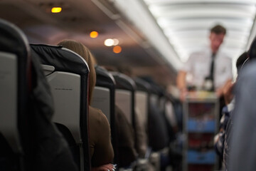 A male steward who distributes food and drinks on board the aircraft. The passengers are sitting in their seats and watching. View along the row. High quality photo