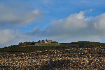Scenic view of a traditional stone house atop a green hill in Normandy, France, with rocky terrain in the foreground and a clear blue sky with scattered clouds.