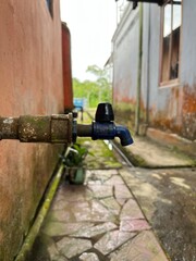 A worn-out blue faucet slowly drips water. The close-up highlights rust, moss, and the gentle flow...