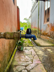 A worn-out blue faucet slowly drips water. The close-up highlights rust, moss, and the gentle flow...