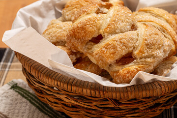 A close-up side view of golden apple strudels arranged in a small basket lined with white paper tissue, placed on a wooden tabletop with a cotton napkin underneath.