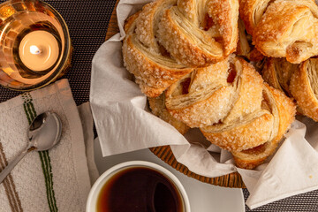 Crispy apple strudels, pastries with a sugar-coated surface arranged in a woven basket lined with white paper, accompanied by a cup of warm tea and a lit candle.