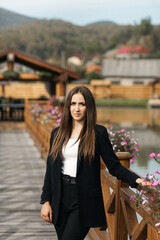 a woman with long brown hair stands on a wooden bridge decorated with flowers. She is wearing a black jacket and a white blouse.