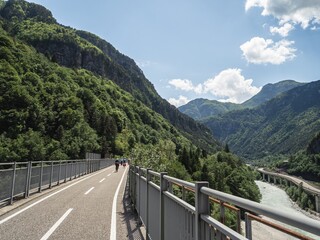 Cyclists ride along the Alpe Adria trail, surrounded by lush greenery and towering mountains under a bright sky with fluffy clouds, creating a perfect outdoor adventure scene.