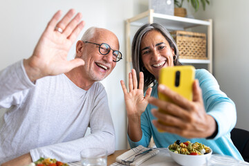 Happy mature married couple waving hand on video call using phone while enjoying meal together at home