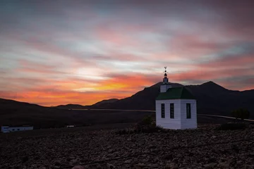 Selbstklebende Fototapeten small cute white wooden chapel in a barren volcanic landscape. Sunrise, landscape shot of Ermita Protestante de Violante church, on Fuerteventura island, Canary Islands, Spain  © Jan