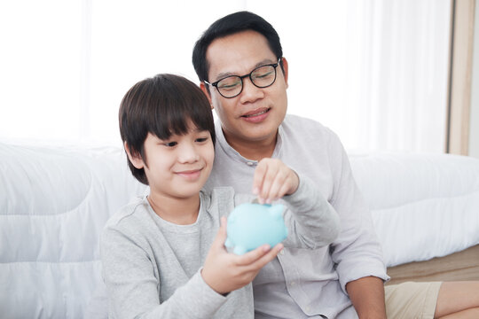 Smiling Asian child boy putting coins into blue piggy bank for saving money with father in bedroom at home. Family love and child educational for homeschool on holiday concept