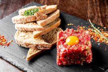 A gourmet beef tartare presented on a slate plate, topped with a raw egg yolk. Accompanied by crispy brown bread slices and garnished with herbs, perfect for fine dining.