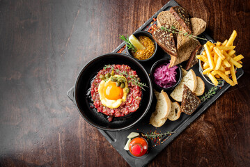 A hearty dish of steak tartare topped with a raw egg yolk, surrounded by garnishes, served with crispy French fries, toasted bread, and pickled onions on a wooden table.