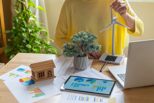 Businesswoman evaluating a miniature wind turbine model, symbolizing investment in renewable energy, surrounded by financial documents, charts, and digital tablet displaying data analysis