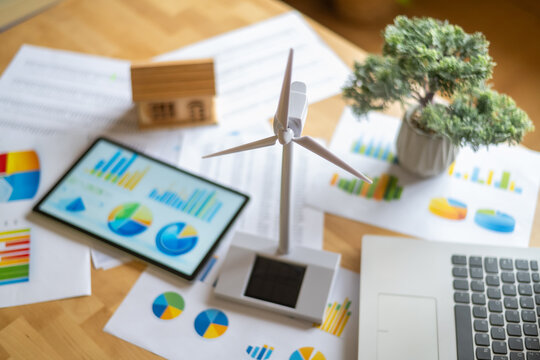 Miniature wind turbine and solar panel models arranged on a desk with financial charts and a digital tablet highlight an engineer assessing renewable energy investments