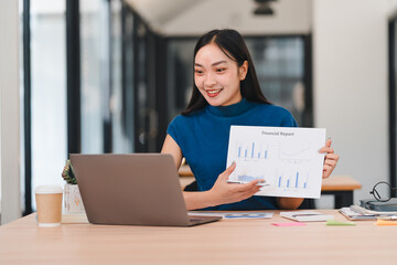 Confident Asian businesswoman in striped shirt and jeans stands in modern office, smiling warmly.