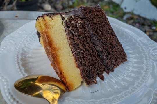 Close-up of a delicious slice of chocolate and vanilla flan cake on an elegant white plate with a golden spoon, taken outdoors. The dessert showcases rich textures and layers.