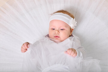 Little baby girl with light hair lying on white fabric, dressed as ballerina. Soft lighting...