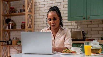 Young african american woman in the morning in the kitchen at home enjoying healthy breakfast, sitting at table working typing on laptop.
