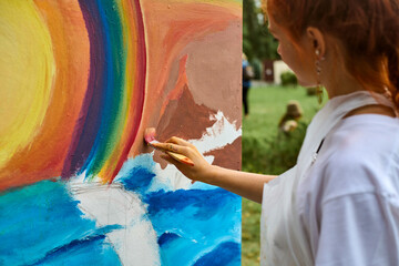 Young female artist with red hair paints vibrant mural in park. Canvas shows large rainbow, ocean landscape. Natural light enhances bright colors and creativity © TRAVELARIUM