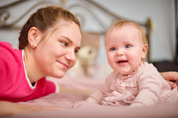 Young mother with brown hair, smiling baby girl indoors. Warm pink tones, soft lighting, metal bedframe in background. Expression of joy and connection