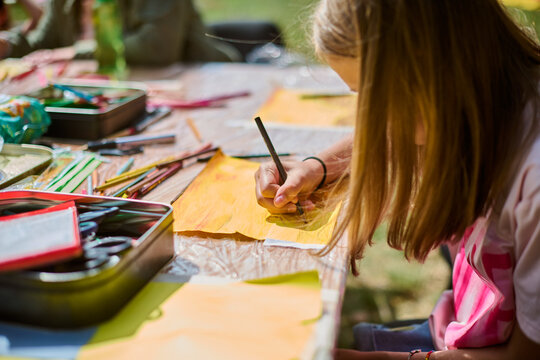 Young child focuses on drawing at outdoor table. Bright natural light, vibrant art supplies, and paper. Creative atmosphere underlines artistic exploration