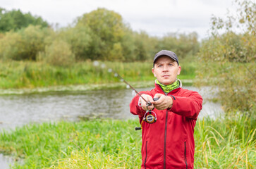 Fisherman in red sweatshirt and cap on the river bank with spinning rod in his hands.