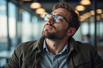 Obraz premium A Relaxed Young Caucasian Man with Glasses Enjoying a Moment of Contemplation in a Modern Office Space with Large Windows and Warm Lighting
