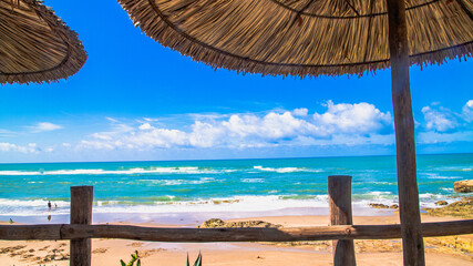 View from a shaded terrace overlooking a paradisiacal beach in Asilah, Morocco. Blue sky, turquoise sea, golden sand, and straw umbrellas create an idyllic atmosphere.